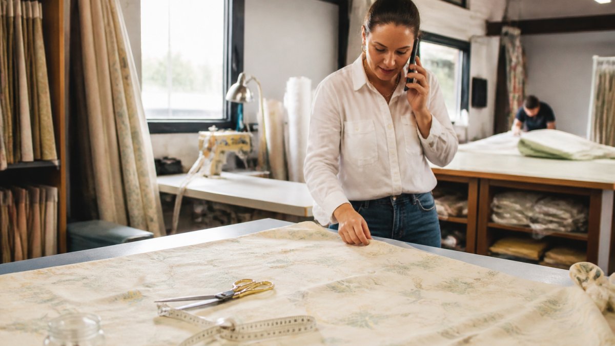 A workroom table with fabric spread out, a measuring tape, scissors, and a sewing machine in the background, showing the hands-on production stage of custom drapery