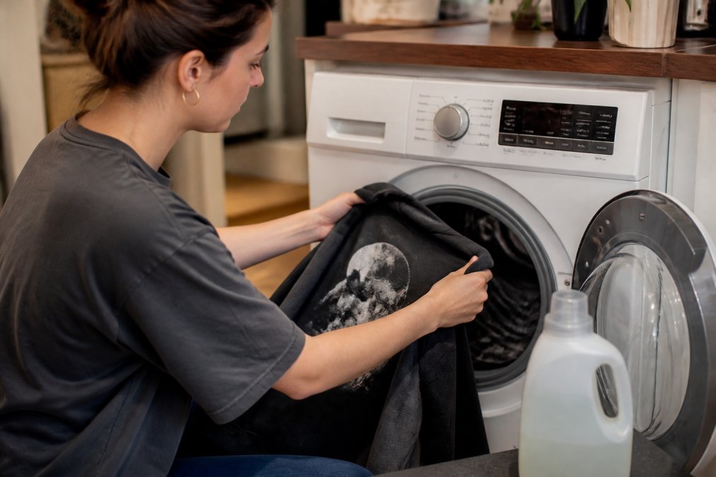 Close-up of hands turning a custom printed shirt inside out before placing it in a washing machine, with a bottle of gentle detergent visible on the counter nearby, natural kitchen lighting