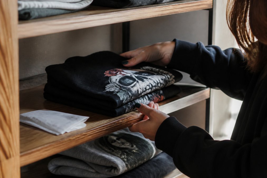 A folded custom printed shirt stored flat on a wooden shelf with a dryer sheet nearby, showing proper storage technique in soft natural light