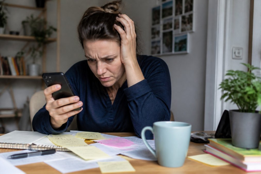 Person sitting at a desk looking frustrated at a smartphone with multiple notification badges visible on the screen, surrounded by scattered notes and a coffee cup