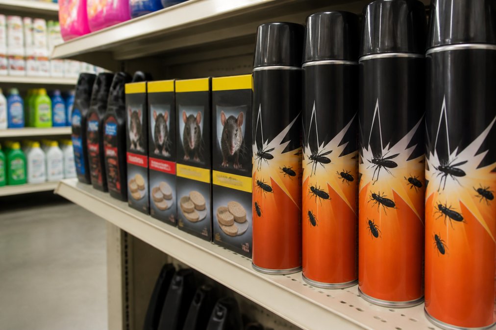 Close-up of a shelf in a hardware shop showing rows of retail pest control sprays and bait boxes in bright fluorescent lighting, with domestic cleaning products visible in the blurred background