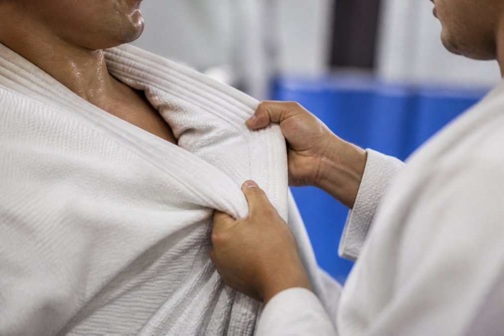 Close-up of a martial artist's hands gripping a white gi lapel during sparring, fabric visibly tensioned and stretched at the collar seam under bright gym lighting