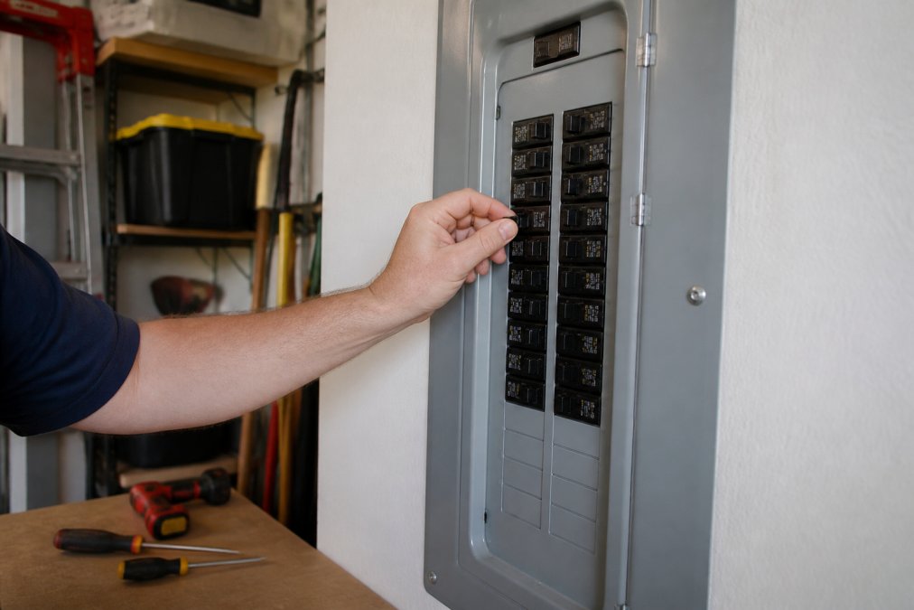 A homeowner's hand reaching toward a residential electrical panel with the breaker switch in the tripped middle position, well-lit garage or utility room setting