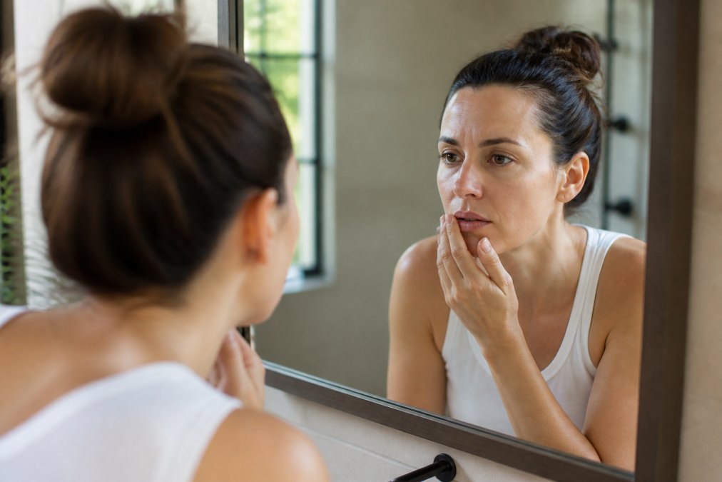 A woman in her 30s examining her lip in a bathroom mirror, touching the corner of her mouth with a concerned expression, soft natural lighting