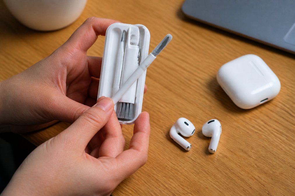 A close-up photo of a person's hands holding a small earbud cleaning kit with a brush and cleaning tool, resting on a wooden desk next to a pair of white wireless earbuds
