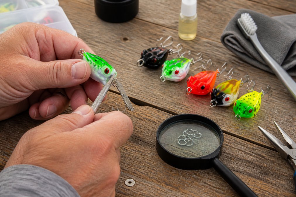 Angler's hands inspecting a fishing lure on a wooden workbench, checking hook sharpness with a file, examining split rings with a magnifying glass, with maintenance tools and cleaning supplies nearby in natural daylight