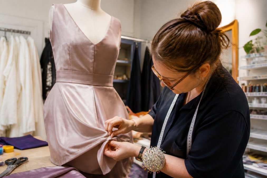 A seamstress carefully pinning the hem of a formal gown on a dress form in a bright tailoring studio, with fabric swatches and measuring tape nearby