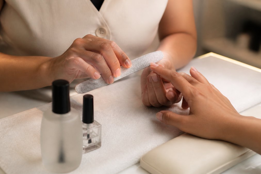 Close-up of a nail technician's hands carefully prepping a client's nails at a clean salon station, with a grit file, dehydrator bottle, and primer visible on the table, under bright warm salon lighting