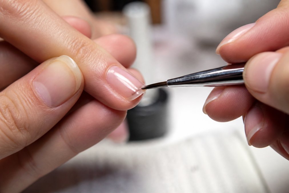 Extreme close-up of a nail technician applying gel polish along the very tip of a client's fingernail, demonstrating the capping technique, with the brush held horizontally against the nail's edge under bright focused lighting