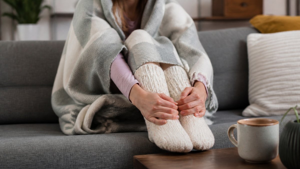 Person sitting on a couch wearing thick warm socks and a blanket, hands wrapping around feet with a warm beverage on a nearby table