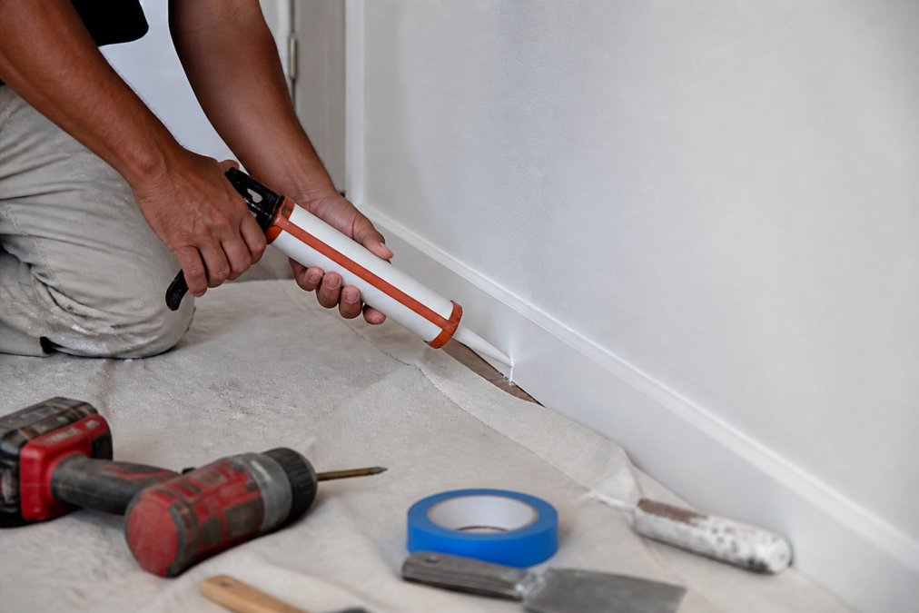 Close-up of hands applying clear caulk to seal a gap between baseboard and wall in a residential apartment, with caulking gun and drop cloth visible