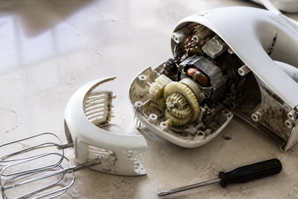 A close-up of a disassembled electric hand mixer on a kitchen counter, showing its worn plastic gears and dusty motor, with a small screwdriver resting nearby under bright task lighting