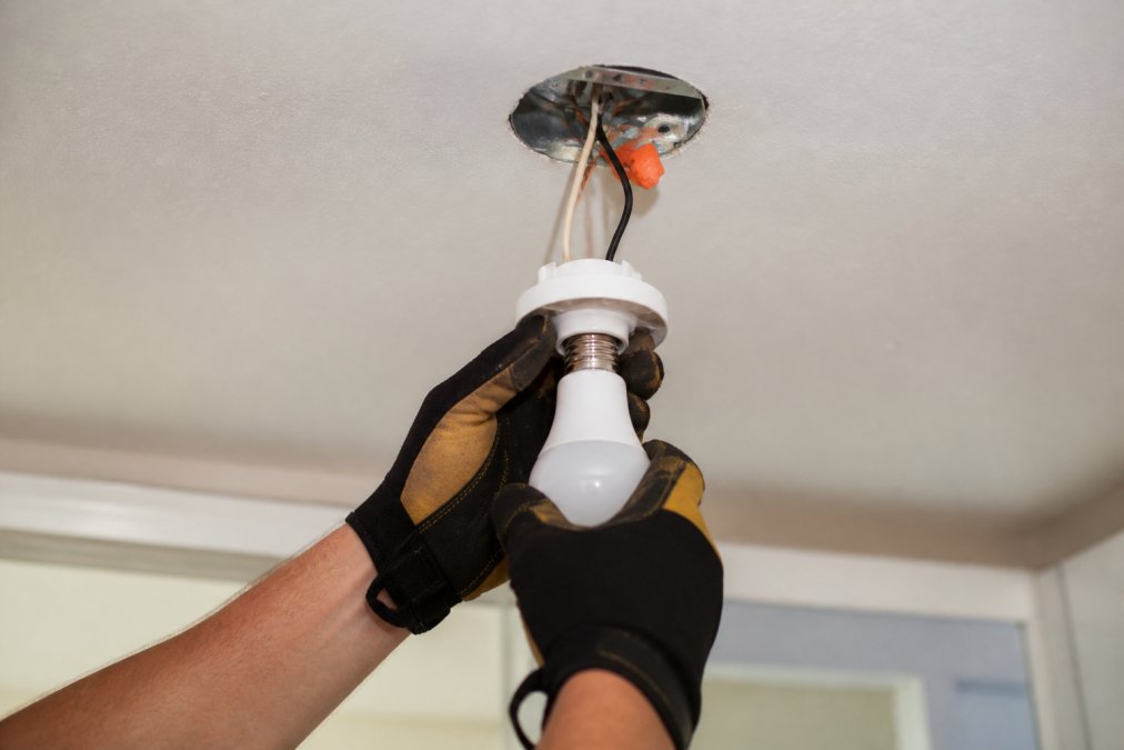 Close-up of an electrician's gloved hands tightening a light bulb into a ceiling fixture, with visible wiring and junction box in the background under bright workshop lighting