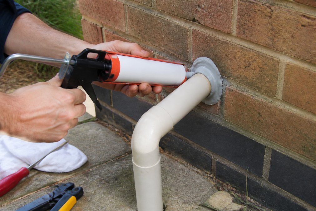 Close-up of hands applying sealant around a pipe where it enters an exterior brick wall, with a tube of sealant and caulking gun visible