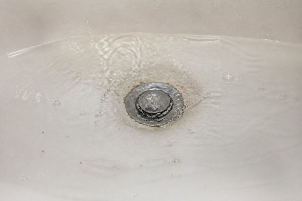 Close-up of water pooling and backing up visibly in a white bathtub drain, with water surface tension visible and subtle discoloration showing debris accumulation