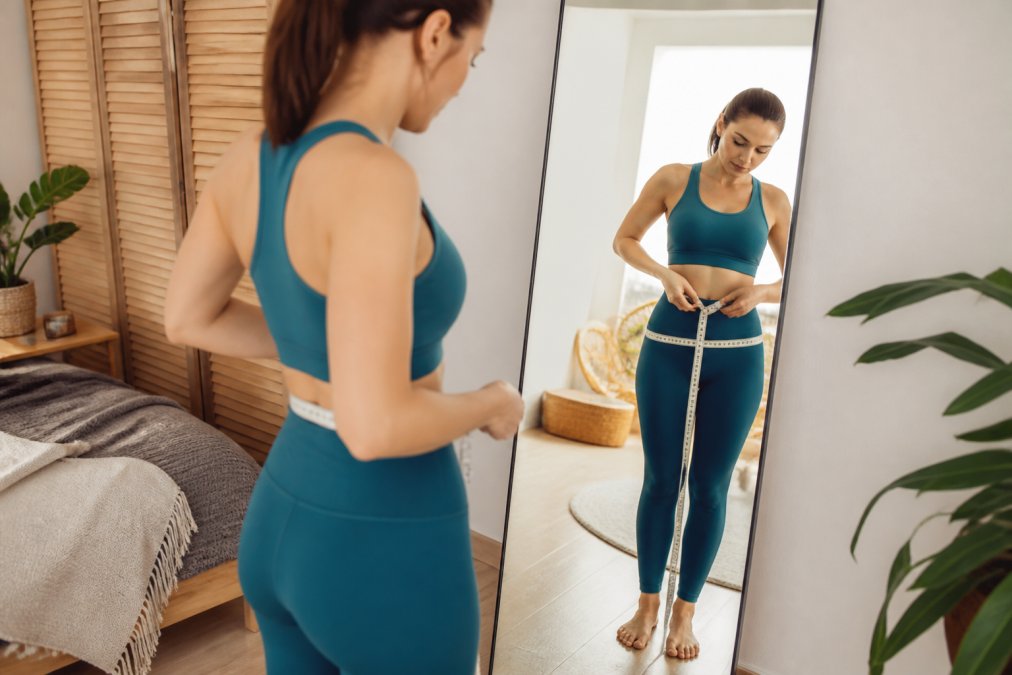 A woman using a soft measuring tape to measure her waist and hips in a bright, clean room with a full-length mirror, showing a natural and practical sizing moment