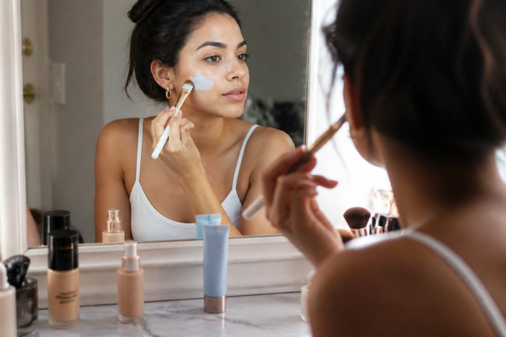 A woman applying a small amount of blue-tinted color correcting primer with a flat brush at a well-lit vanity mirror, with neatly arranged makeup products including foundation bottles and a primer on the marble surface in front of her