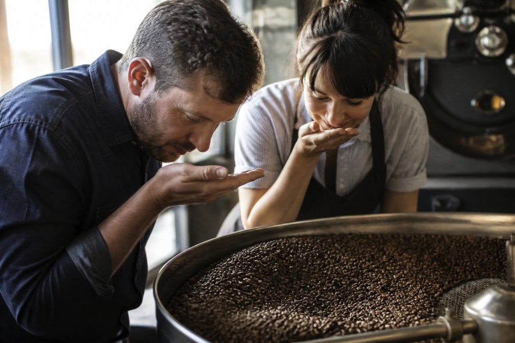 Close-up of freshly roasted coffee beans in a ceramic bowl, visible oils on the bean surface, warm natural window light, faint steam rising, wooden countertop
