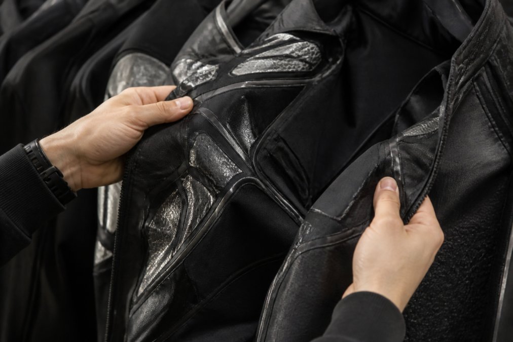 Close-up of hands holding a futuristic black technical jacket with reflective silver panels and intricate seam work, showing detailed craftsmanship and premium fabric texture under soft studio lighting
