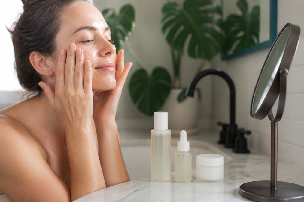 Hands applying a hydrating serum to face with visible moisture, with skincare products (toner, moisturizer, and serum bottles) arranged on a clean bathroom counter with natural lighting