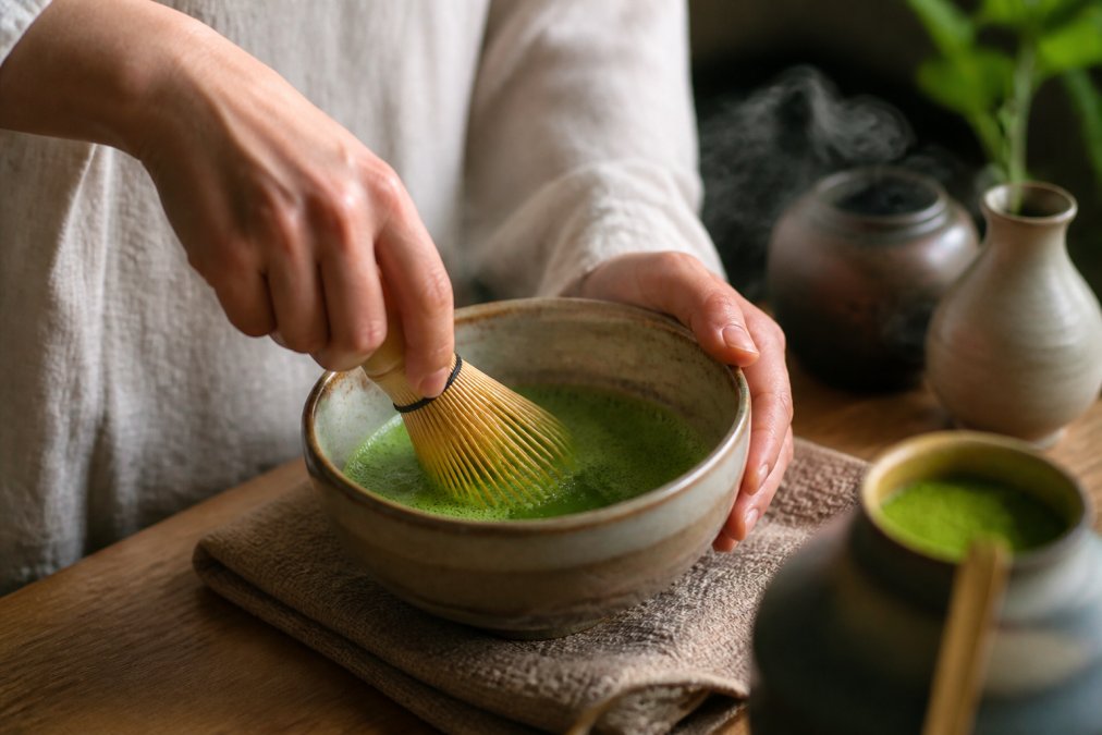 Close-up of hands holding a bamboo whisk whisking vibrant green matcha in a ceramic bowl, soft natural light, steam rising gently