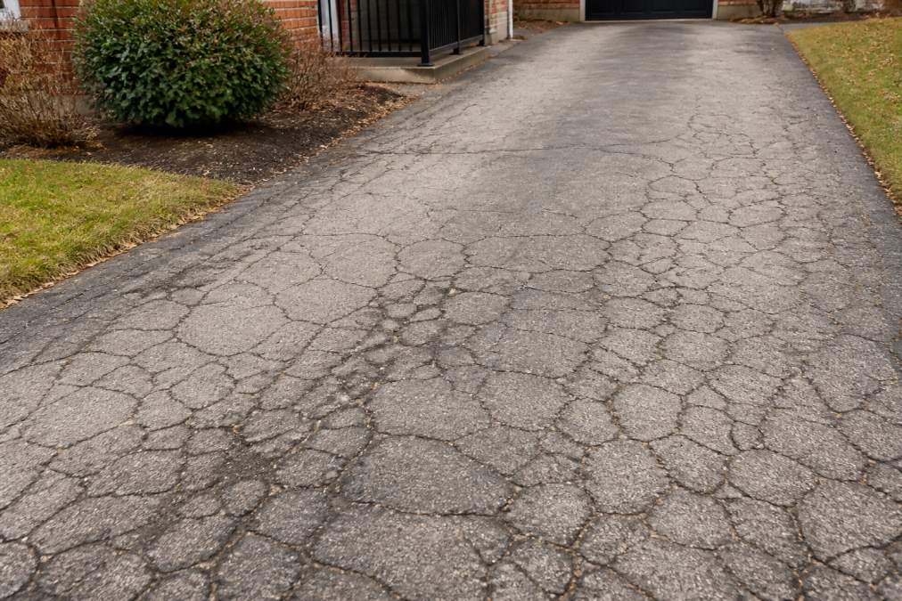 Residential driveway with visible alligator cracking pattern, dark surface fractures spreading across pale asphalt, showing progressive deterioration and uneven settling damage