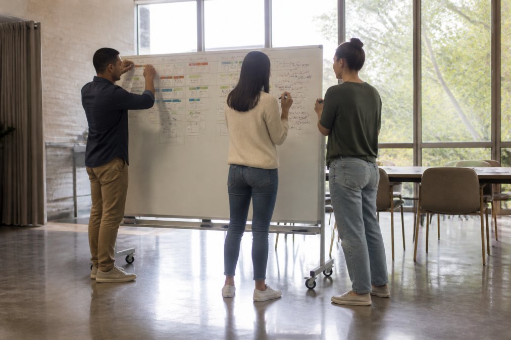 A diverse product team in a bright, modern office gathers around a large freestanding magnetic whiteboard on wheels, one person moving a task card while two others write notes, natural daylight flooding the space
