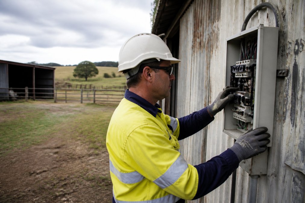 A licensed electrician in safety gear inspecting a rural property switchboard mounted on a weathered exterior wall, with farm sheds and paddocks visible in the background under an overcast sky