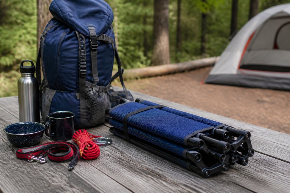 Compact folded elevated dog bed in navy mesh next to a hiking backpack and camping essentials on a picnic table, demonstrating how portable gear integrates into camping setup
