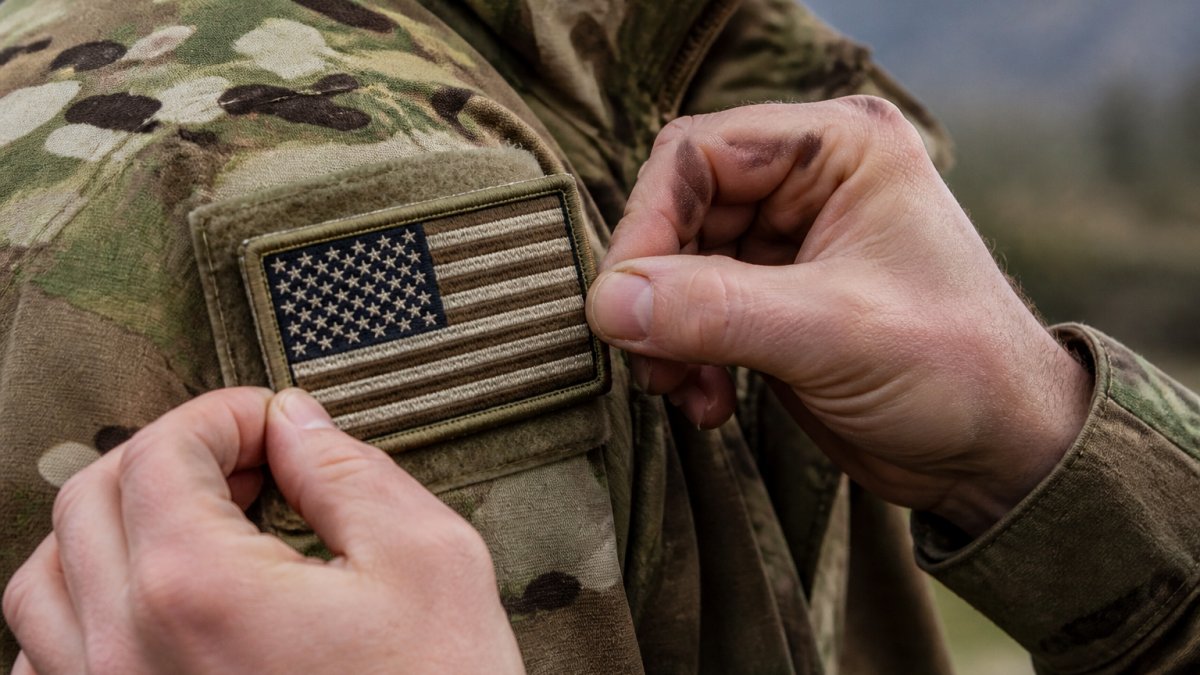 Close-up of an embroidered military patch on a tactical jacket, showing detailed stitching and natural thread against durable fabric, with a service member's hands adjusting the patch in an outdoor field setting