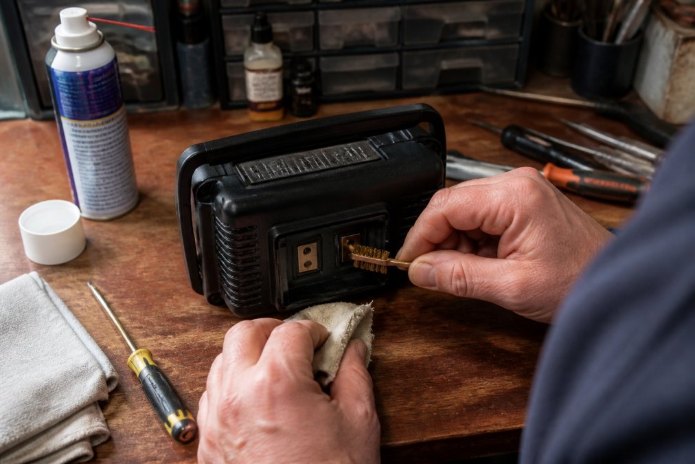 Person carefully cleaning radio battery terminals with a small brass brush and cloth on a workbench with various tools and supplies nearby, under bright workshop lighting