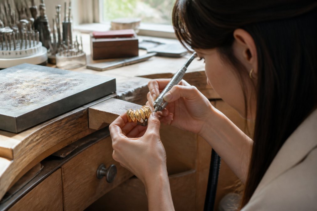 Skilled artisan jeweler working at a wooden bench in a bright, naturally-lit workshop, hands carefully crafting a gold jewelry piece with precision metalworking tools, showing the detailed labor involved in ethical jewelry production
