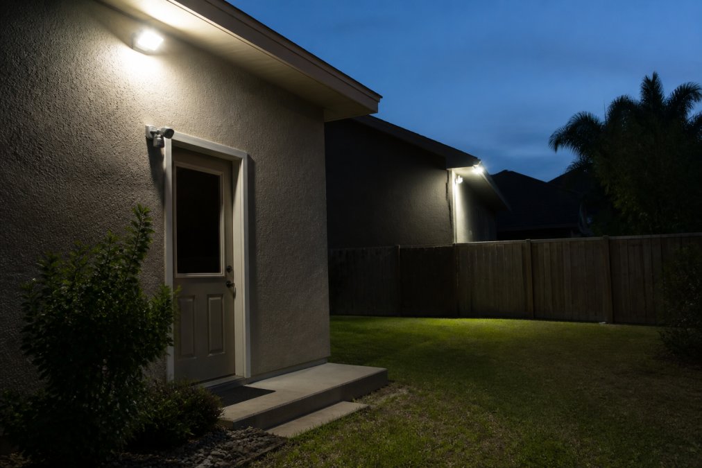 Exterior home at dusk showing motion-activated floodlights mounted on corners illuminating a dark backyard with sensor visible near an entry point, creating bright zones around the perimeter