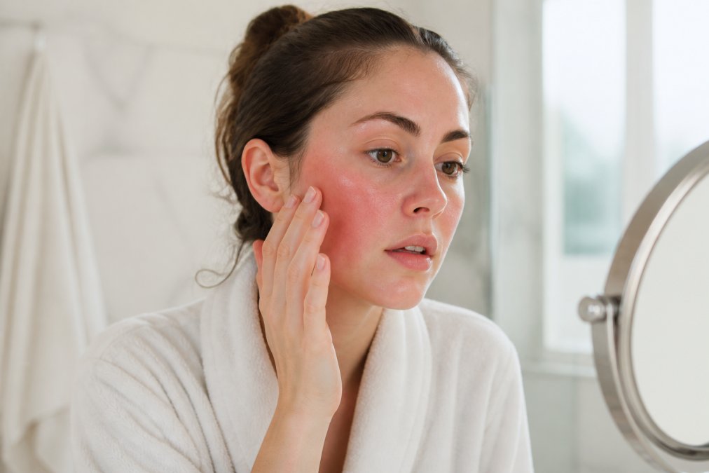 A woman gently touching her dry, slightly flushed cheek after washing her face in a bright, clean bathroom with soft natural light