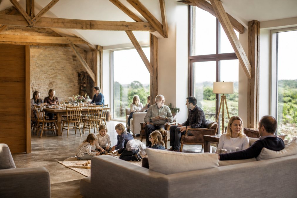 Multi-generational family gathered in a spacious countryside barn interior, with children playing on the floor, adults chatting in comfortable seating areas, and natural light streaming through large windows