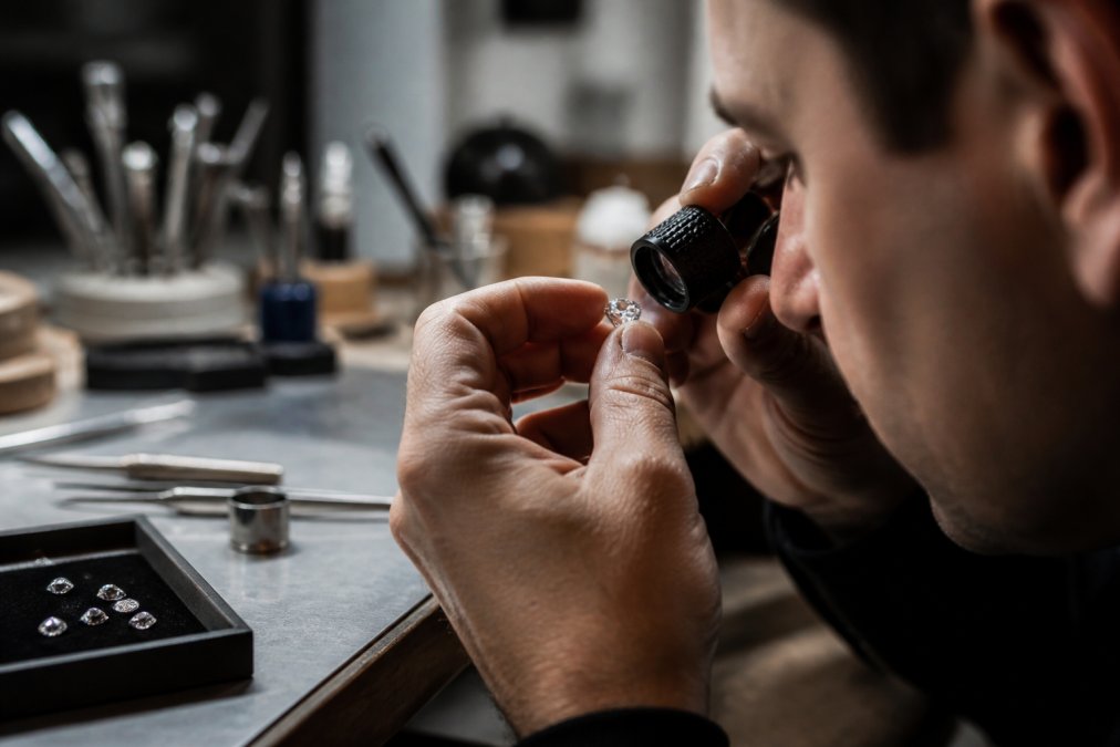 Jeweller's loupe held in steady hands examining a diamond under bright task lighting, gemstone reflecting light, professional workbench with jewellery tools visible in soft focus