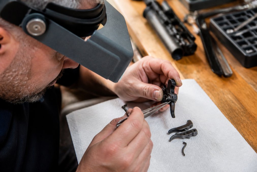 Gunsmith examining internal trigger assembly components including sear, disconnector, and hammer under magnification with precision tools on workbench
