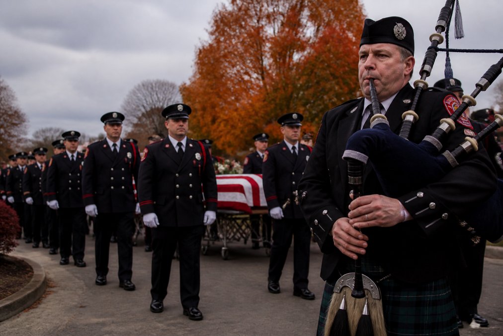 Bagpiper in dress uniform with Great Highland Bagpipes playing at firefighter memorial service, with honor guard in dark dress uniforms and firefighters in solemn procession behind, autumn trees and gray sky in background