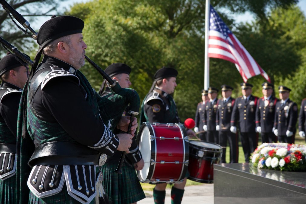 Fire department pipe and drum band in full ceremonial dress uniforms with Great Highland Bagpipes and drums performing at outdoor memorial service, with firefighters in dress blues standing at attention in background, American flag visible