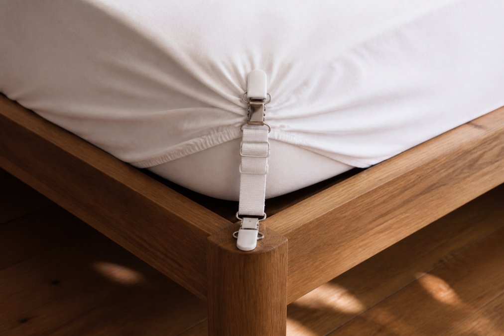 Close-up of fitted sheet corner clips gripping the elastic corner of a white sheet, anchored to a wooden bed frame with natural morning light