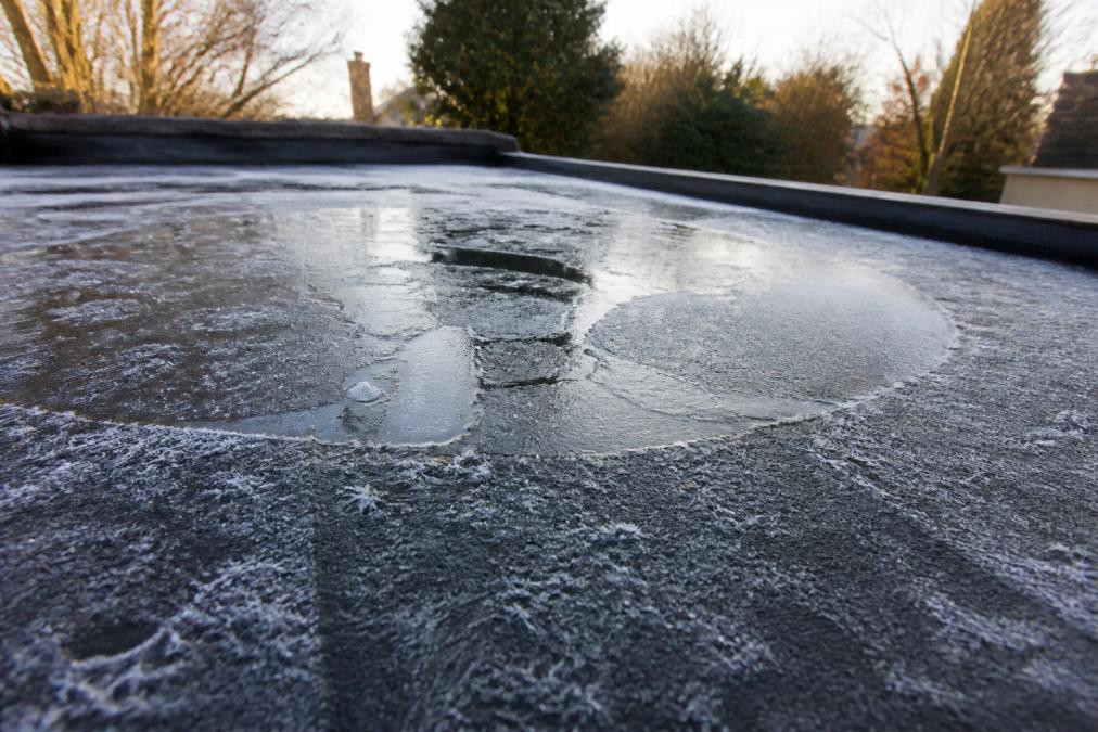 Close-up of flat roof surface showing ice formation, frost patterns, and water pooling during winter conditions with morning frost glistening