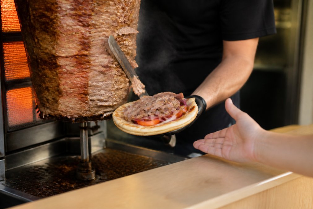Close-up of vertical rotisserie with freshly sliced gyro meat falling onto warm pita bread, steam rising, with customer's hand reaching for it in warm golden lighting