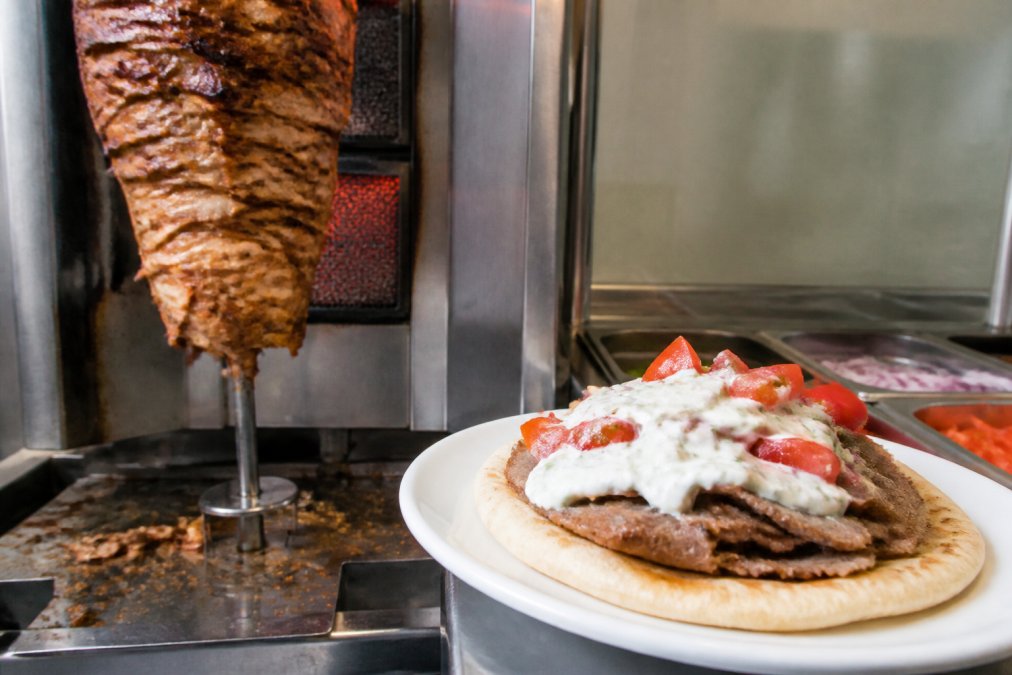 Side-by-side scene of freshly prepared gyro with charred, crispy edges on vertical rotisserie next to finished gyro sandwich topped with fresh tomatoes, onions, and tzatziki sauce on white plate
