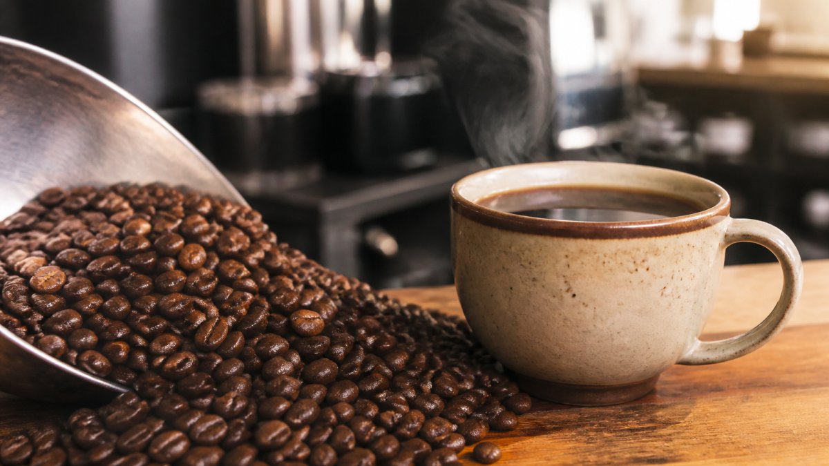 Close-up of freshly roasted coffee beans with visible oils, next to a ceramic cup of brewed coffee on a wooden counter, steam rising, warm natural light