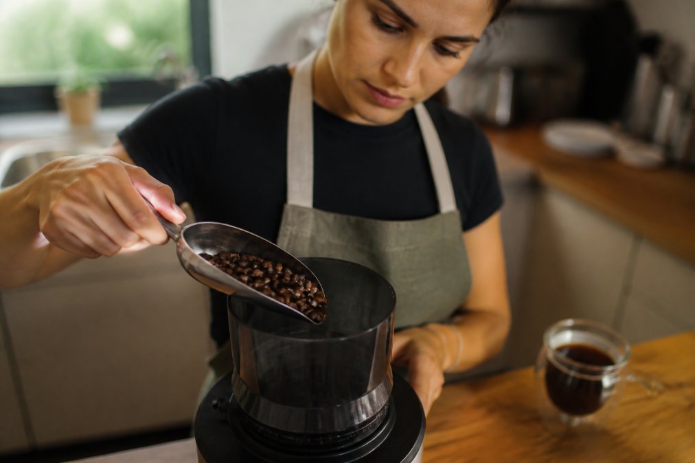 Close-up of a hand holding a scoop of freshly roasted coffee beans above a burr grinder, with a glass of brewed coffee blurred in the background, warm kitchen lighting