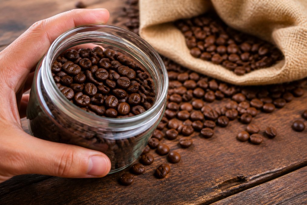 Close-up of a hand holding a clear glass jar filled with freshly roasted coffee beans, glossy surface visible under warm light, with loose beans scattered on a rustic wooden table beside a burlap coffee sack