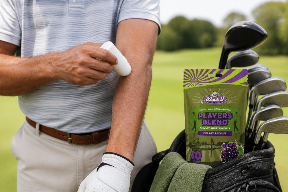 Close-up of a golfer's hands applying a white therapy stick to his forearm near a golf bag, with a green golf course visible in the soft-focus background
