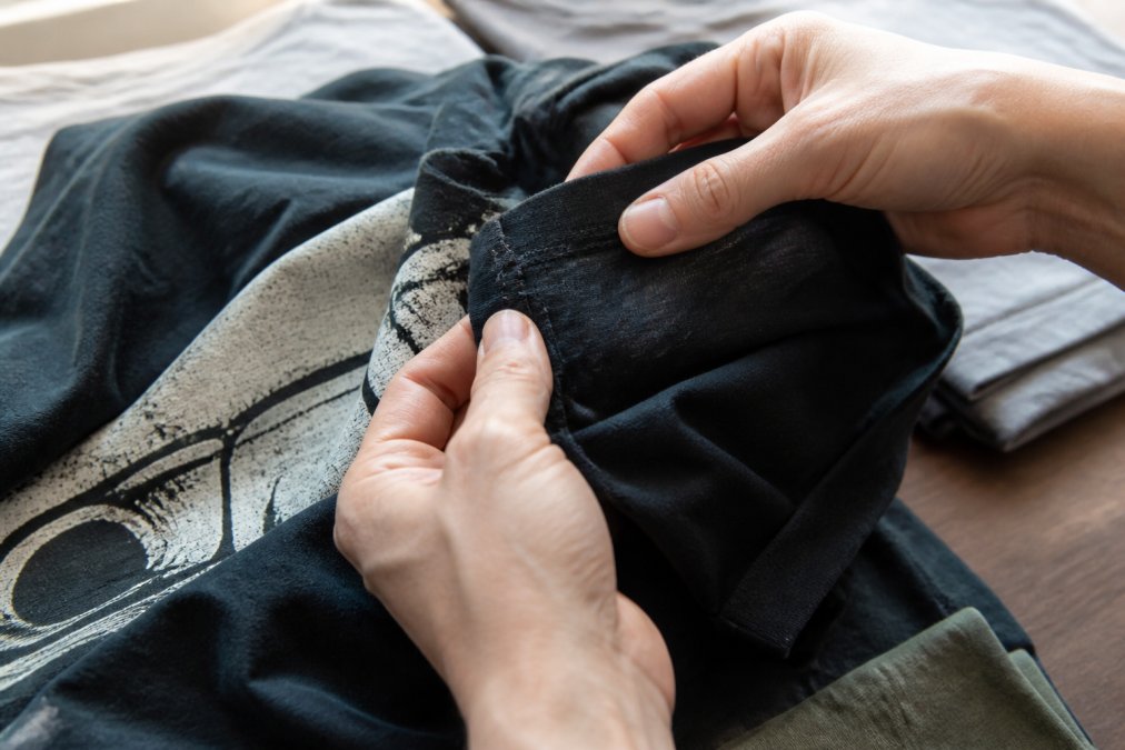 Close-up of hands examining a graphic tee's fabric texture, seams, and print quality under natural light, showing detailed inspection of stitching and ink durability