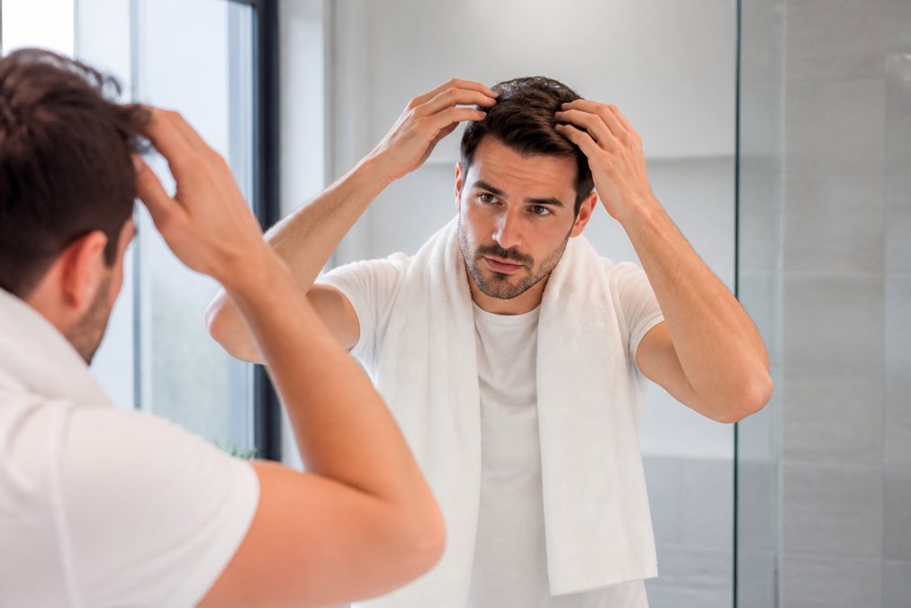 A man in his thirties in a bright bathroom mirror, applying styling gel evenly through damp hair with his fingers, focused expression, natural light, no visible white residue on hair