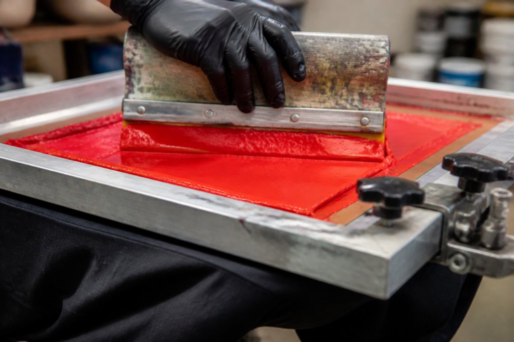 A gloved hand pulling a squeegee across a screen printing press, forcing a thick layer of plastisol ink through the mesh into a t-shirt fabric below
